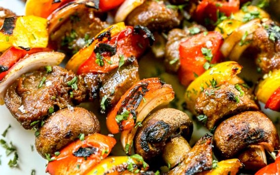 Grilled steak kabobs with peppers, onions, and mushrooms on a white plate, sprinkled with herbs.