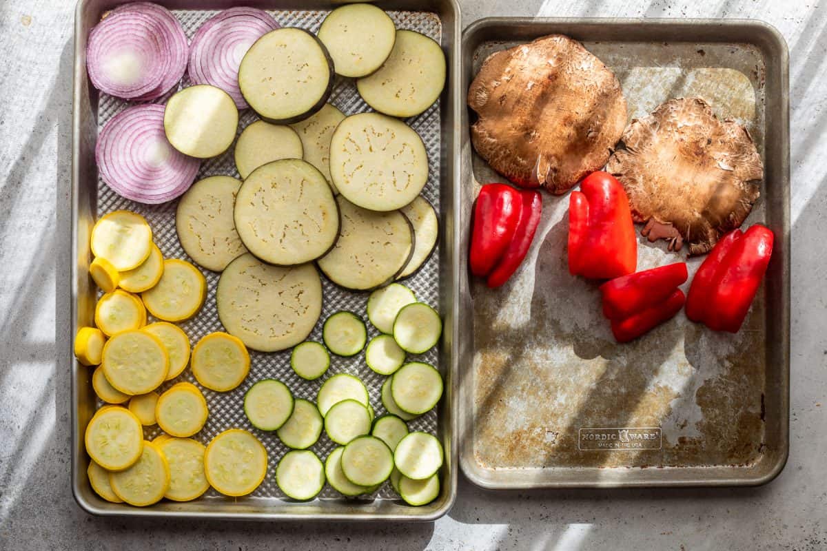 Sliced onions, eggplant, yellow squash, and zucchini on one tray; portobello mushrooms and red bell peppers on another.