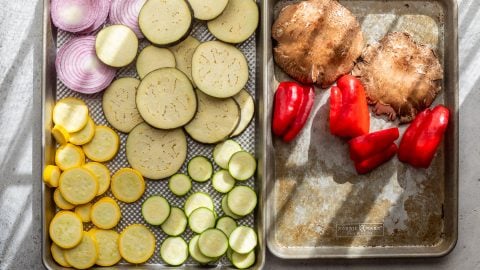 Sliced onions, eggplant, yellow squash, and zucchini on one tray; portobello mushrooms and red bell peppers on another.