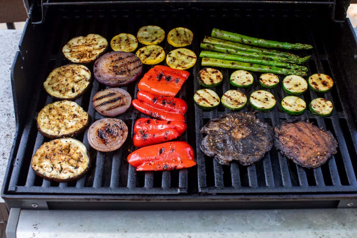 Assorted vegetables, including mushrooms, onions, peppers, zucchini, squash, and asparagus, grilling on a barbecue.