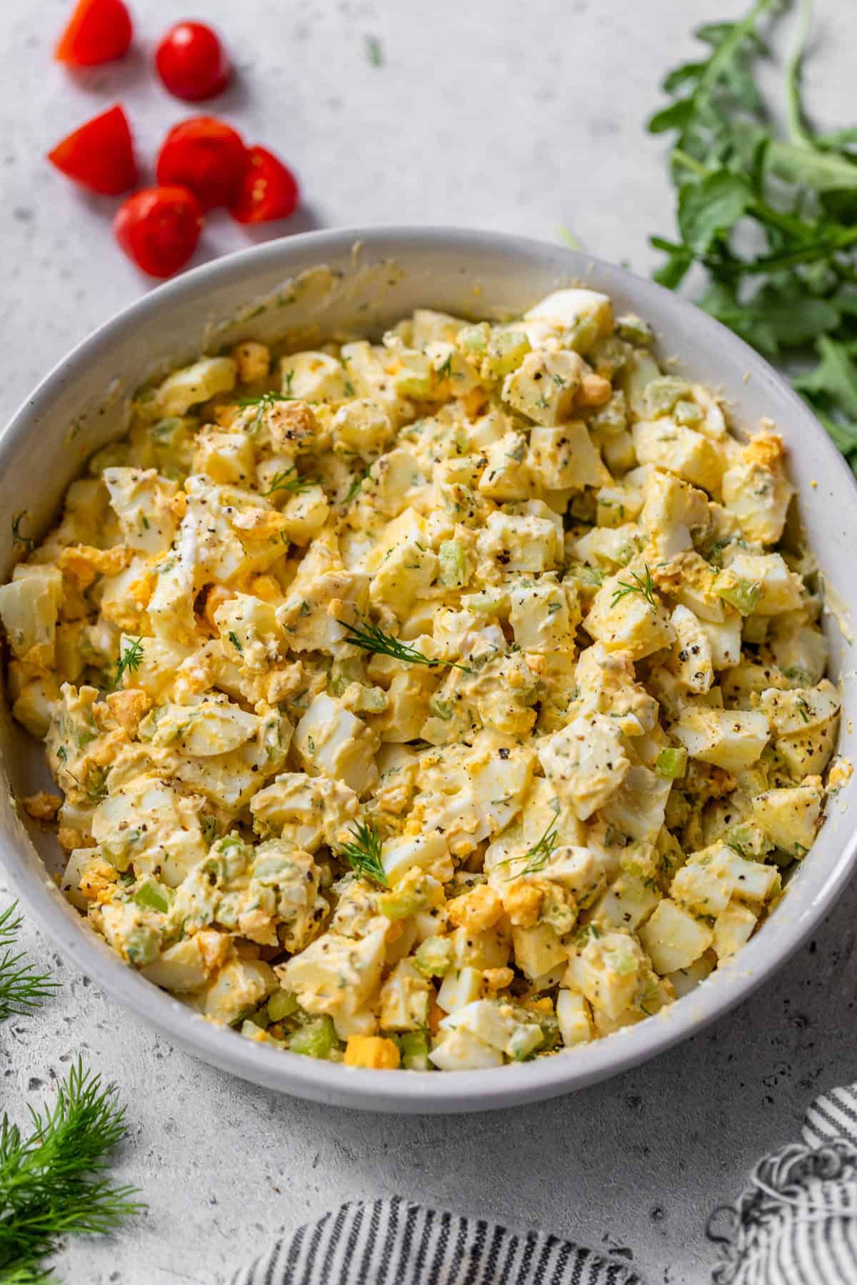 A bowl of healthy egg salad with herbs, surrounded by fresh dill, parsley, and sliced cherry tomatoes on a light surface.