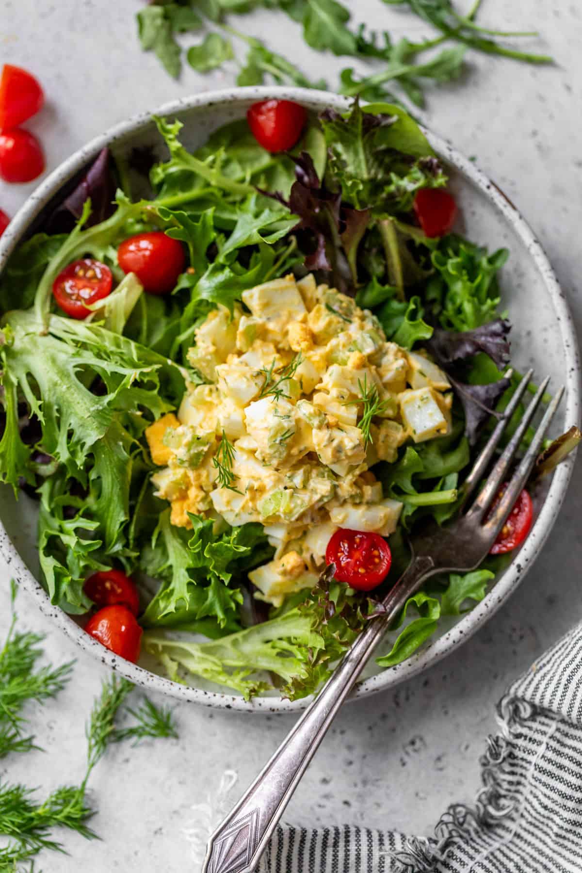 A bowl of mixed greens topped with healthy egg salad and cherry tomato halves, with a fork resting on the side.
