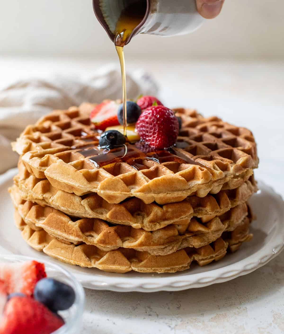 A stack of healthy waffles topped with berries is drizzled with syrup from a small pitcher.