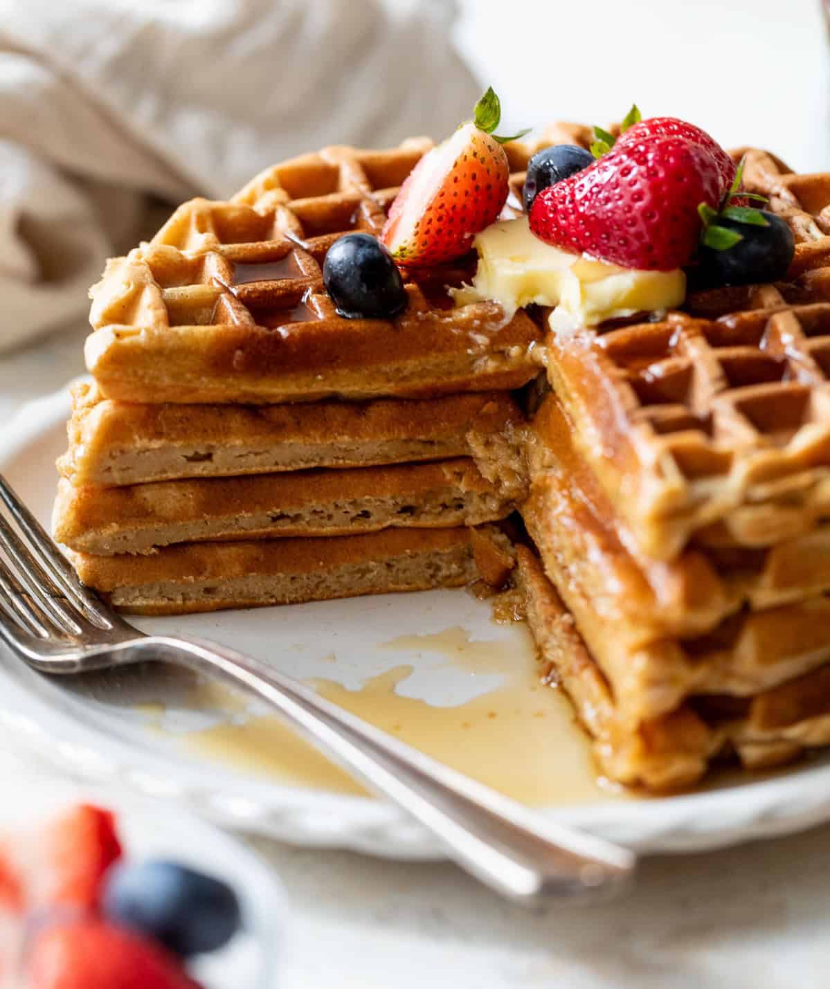 A stack of healthy waffles topped with butter, strawberries, blueberries, and syrup, with a slice cut out on a white plate.