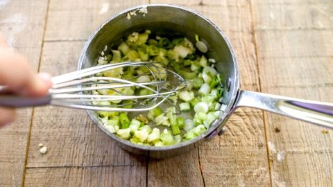 A hand whisking chopped green onions and garlic in a small saucepan on a wooden surface.
