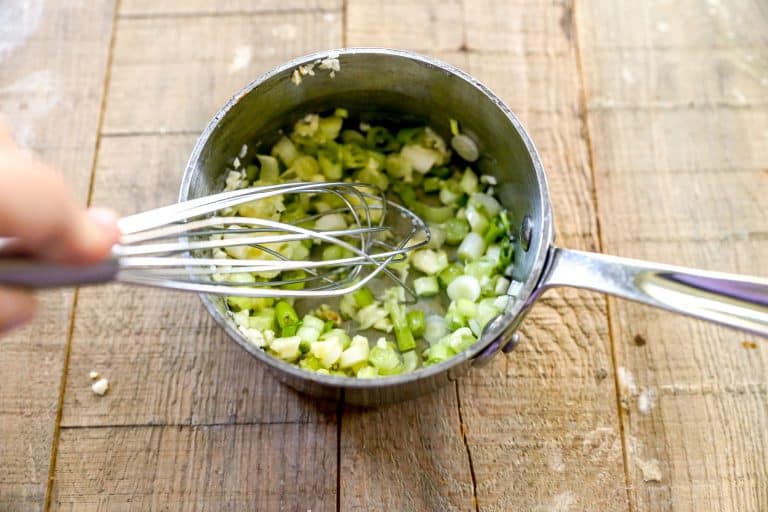 A hand whisking chopped green onions and garlic in a small saucepan on a wooden surface.