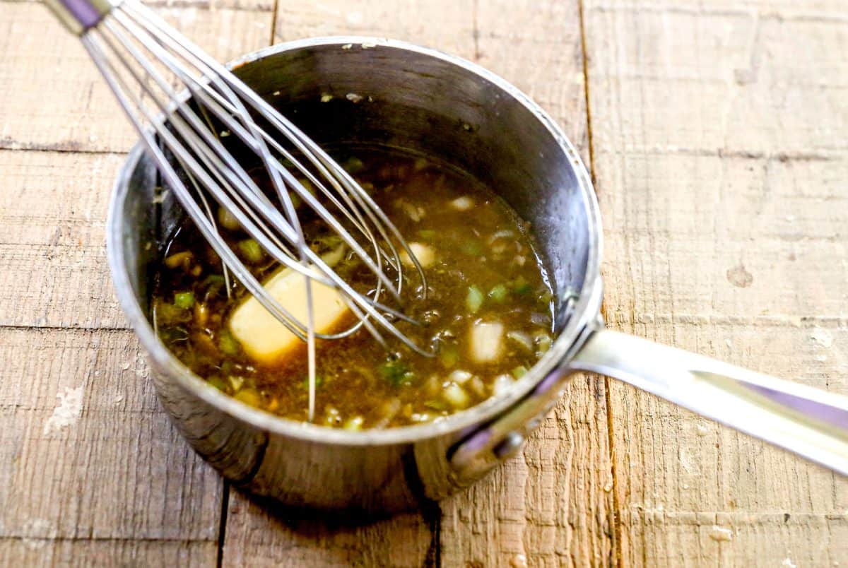 A whisk stirring sauce with butter and chopped onions in a small saucepan on a wooden surface.