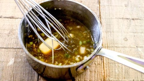 A whisk stirring sauce with butter and chopped onions in a small saucepan on a wooden surface.