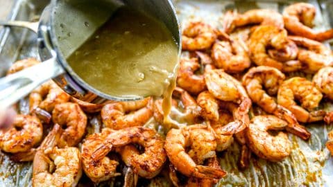 A hand pours sauce from a small pot over seasoned BBQ shrimp on a baking sheet.