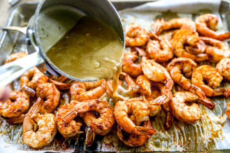 A hand pours sauce from a small pot over seasoned BBQ shrimp on a baking sheet.