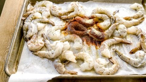 Raw shrimp arranged on a parchment-lined baking sheet with spices sprinkled in the center.