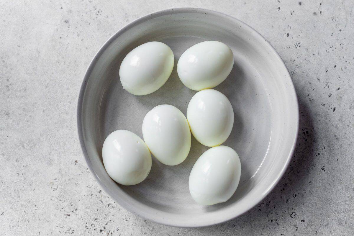 Six peeled hard-boiled eggs arranged in a white bowl on a light gray speckled surface.