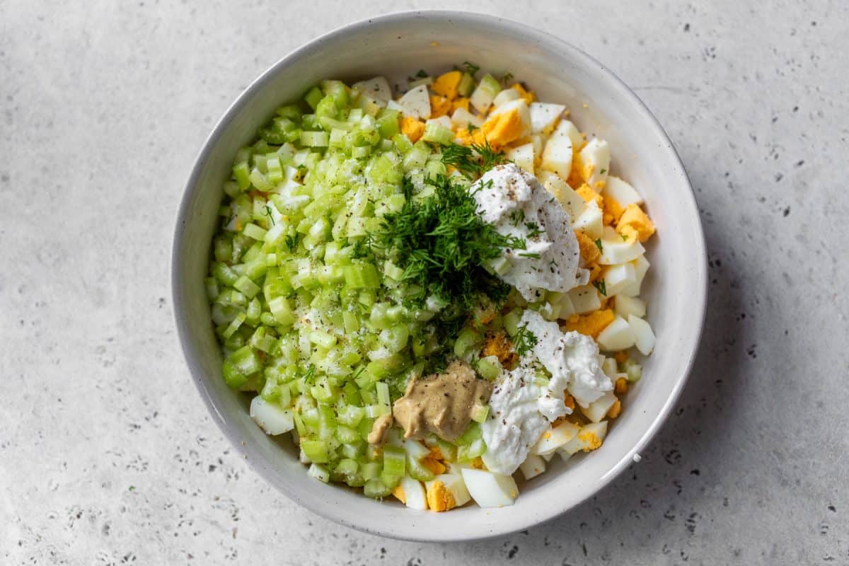 A bowl with chopped celery, hard-boiled eggs, fresh dill, mustard, and dollops of yogurt or mayonnaise on a light surface.