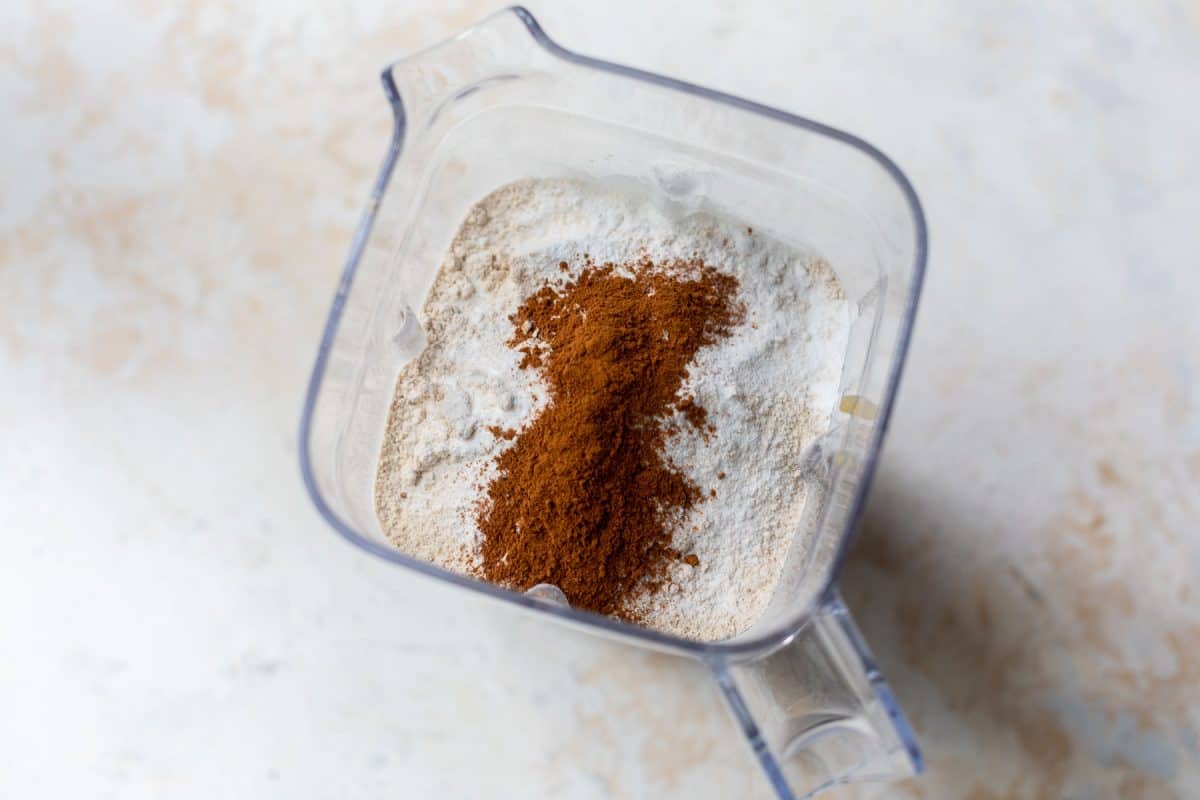 Top view of a blender containing flour and a pile of ground cinnamon on a light surface.