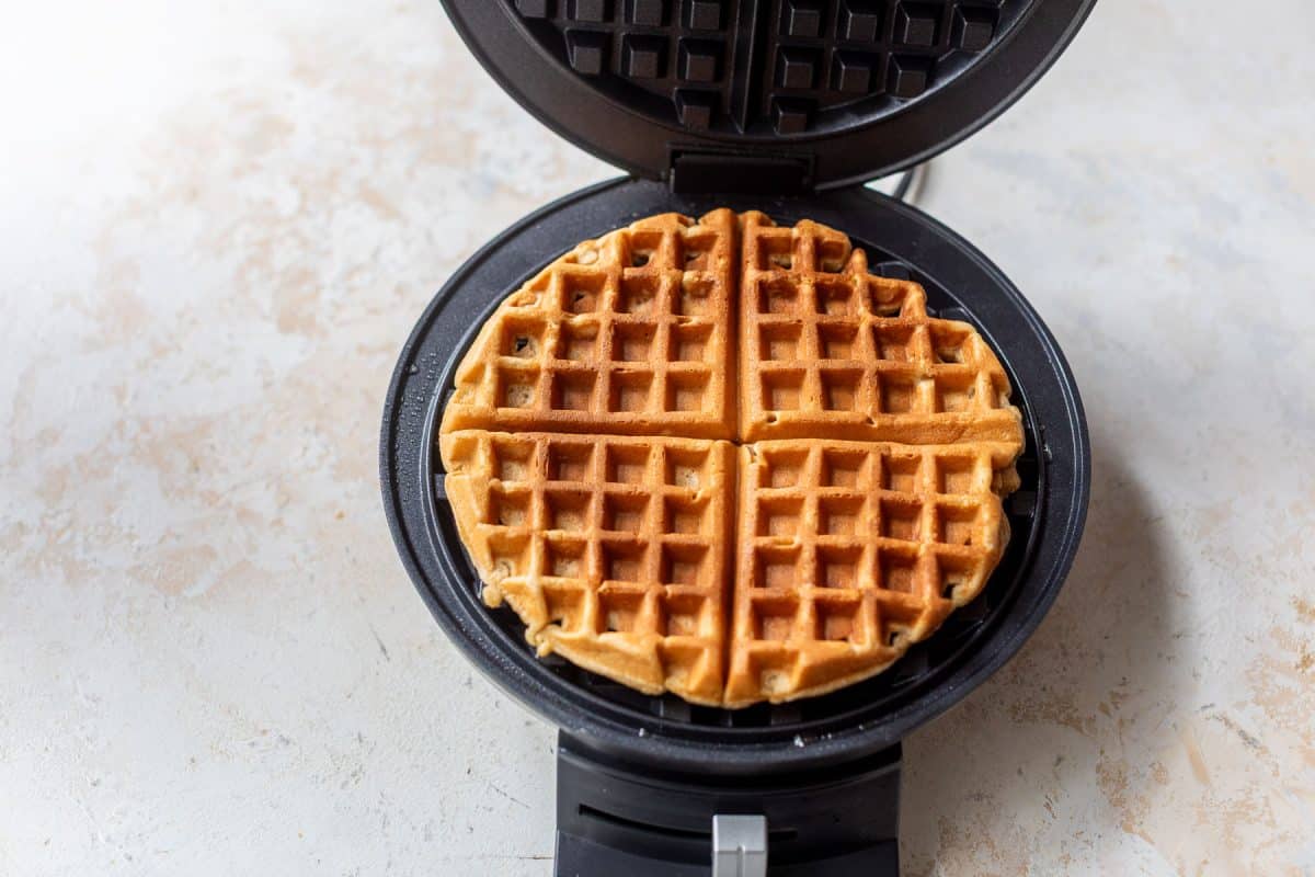 A golden-brown healthy waffle in a black waffle maker on a light-colored countertop.