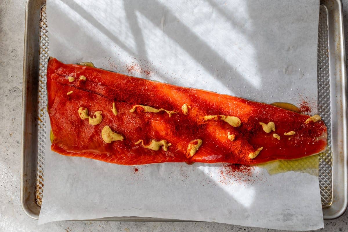 A raw salmon fillet on parchment paper, topped with mustard and garlic, on a baking sheet.