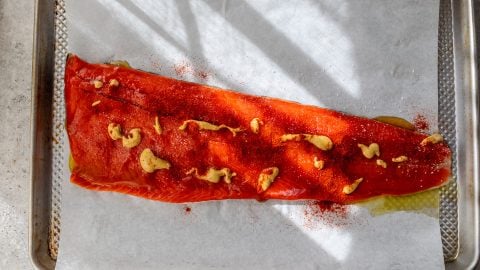 A raw salmon fillet on parchment paper, topped with mustard and garlic, on a baking sheet.