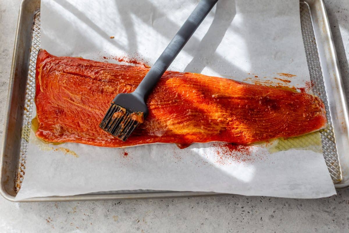A brush spreads honey mustard on a raw salmon fillet placed on parchment paper on a baking sheet.
