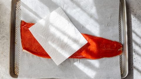 A raw salmon fillet on parchment paper with a paper towel on top, on a baking sheet in sunlight.