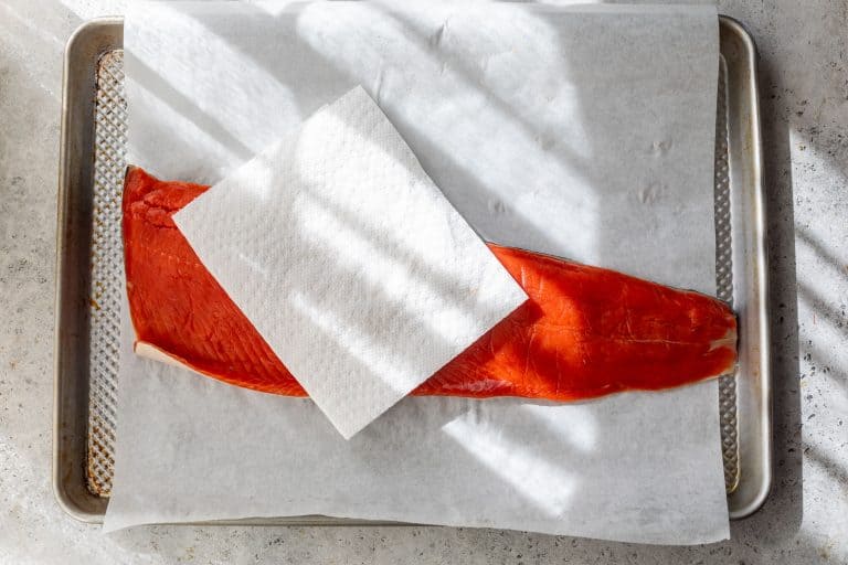 A raw salmon fillet on parchment paper with a paper towel on top, on a baking sheet in sunlight.