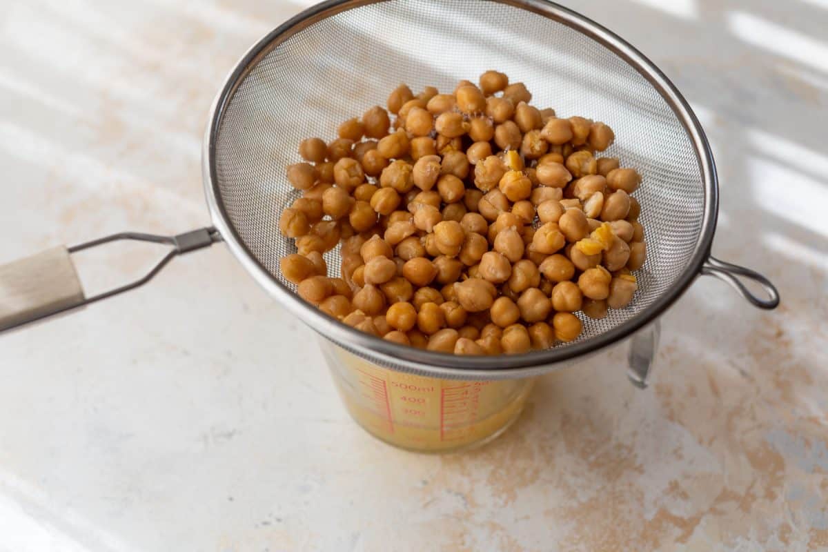Chickpeas draining in a metal strainer over a glass measuring cup on a light-colored surface.