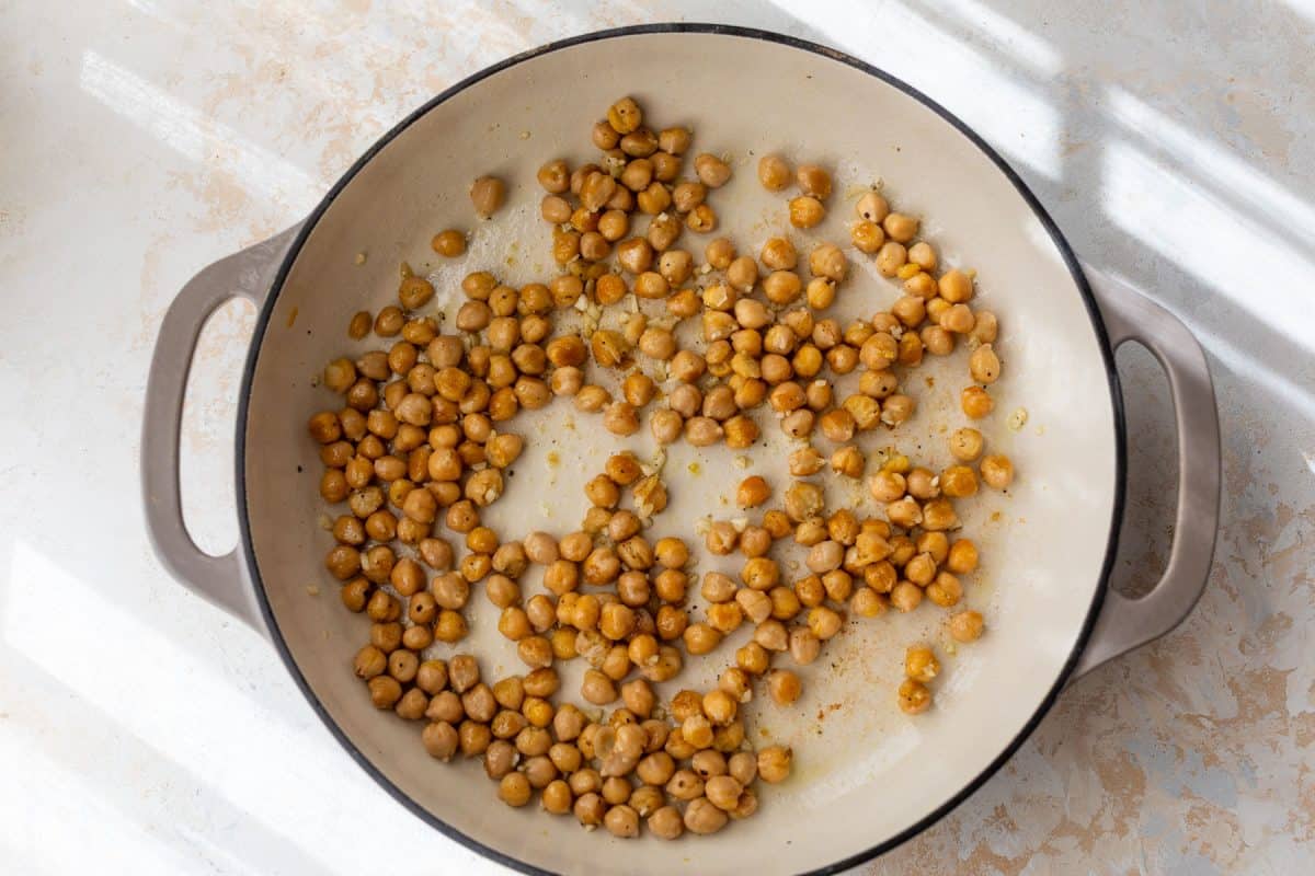 Chickpeas being sautéed in a large beige skillet on a light-colored surface, lit by natural sunlight.