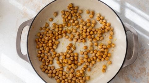 Chickpeas being sautรฉed in a large beige skillet on a light-colored surface, lit by natural sunlight.
