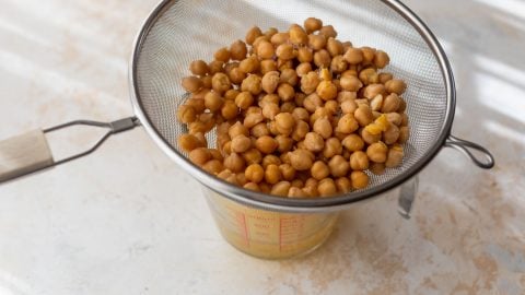 Chickpeas draining in a metal strainer over a glass measuring cup on a light-colored surface.