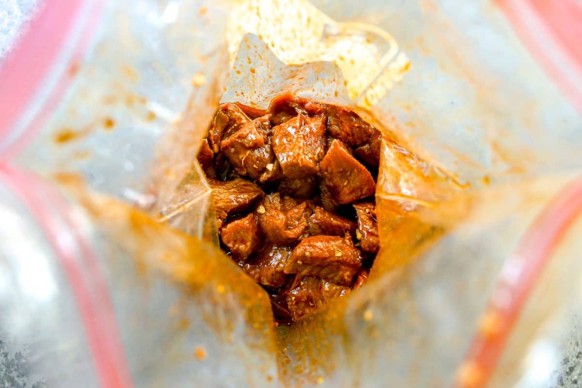 Chunks of raw steak marinating in a reddish sauce inside a clear plastic zip-top bag.