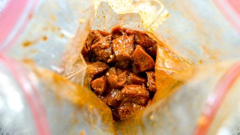 Chunks of raw steak marinating in a reddish sauce inside a clear plastic zip-top bag.