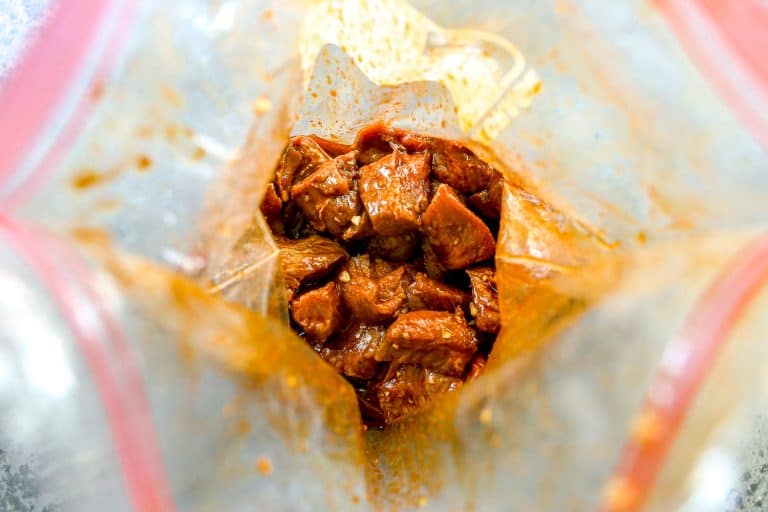 Chunks of raw steak marinating in a reddish sauce inside a clear plastic zip-top bag.