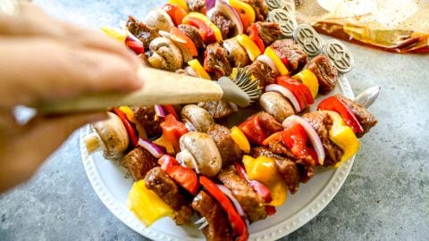 A hand places grilled vegetable and steak skewers with peppers, onions, and mushrooms on a white plate.