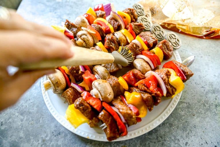 A hand places grilled vegetable and steak skewers with peppers, onions, and mushrooms on a white plate.