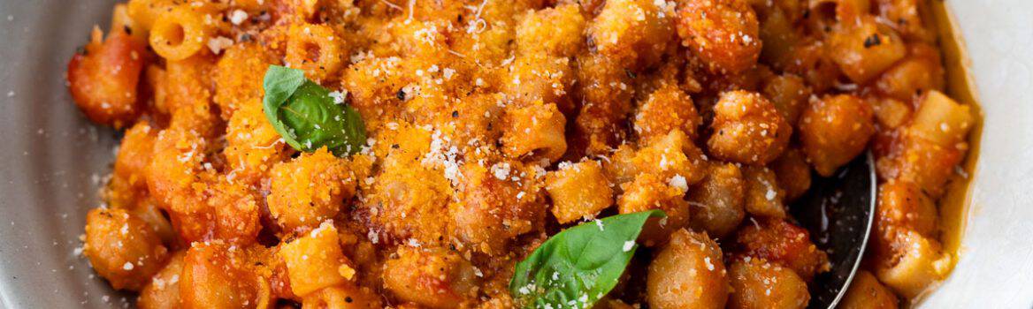 A bowl of pasta e ceci in tomato sauce, topped with grated cheese and basil leaves, with a spoon in the bowl.
