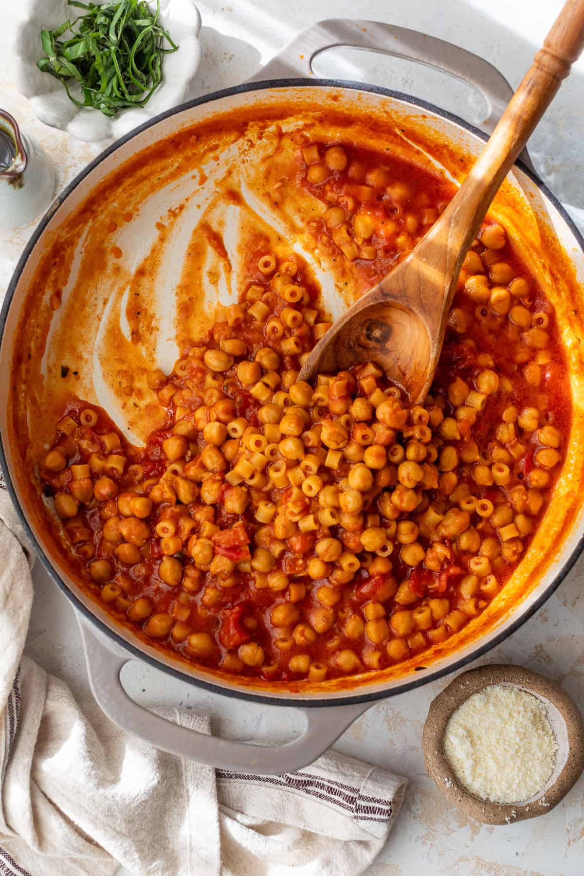 A pot of pasta with chickpeas in tomato sauce, stirred with a wooden spoon, beside spinach and grated cheese.