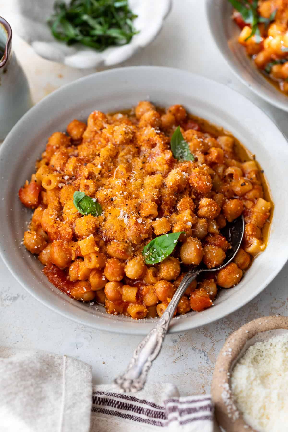 A bowl of pasta e ceci in tomato sauce, topped with grated cheese and basil leaves, with a spoon in the bowl.