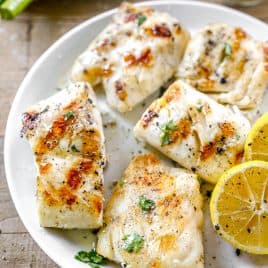 Grilled cod fillets on a plate with lemon slices, herbs, and a side of sauce.