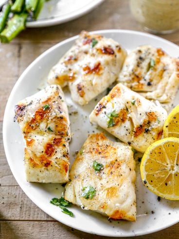 Grilled cod fillets on a plate with lemon slices, herbs, and a side of sauce.