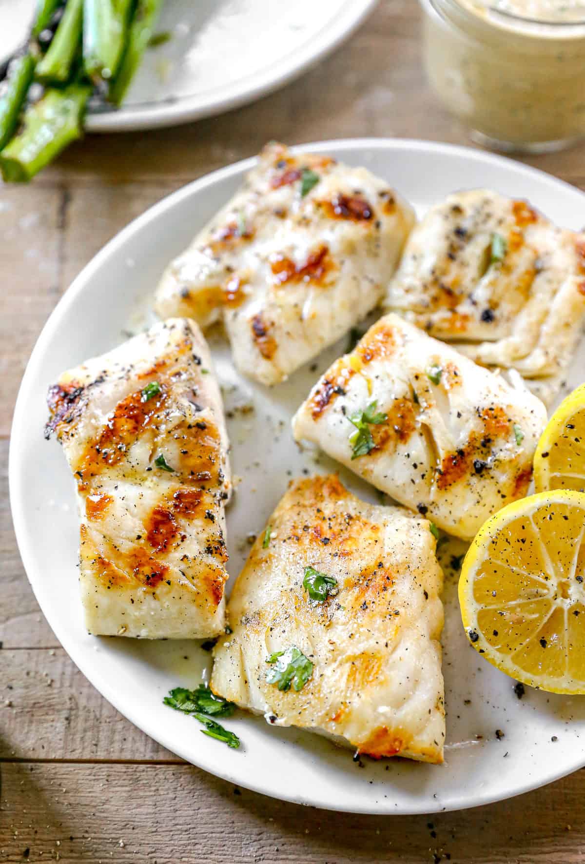Grilled cod fillets on a plate with lemon slices, herbs, and a side of sauce.