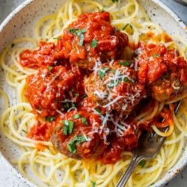 A bowl of spaghetti topped with ground beef meatballs, tomato sauce, grated cheese, and chopped herbs with a fork.