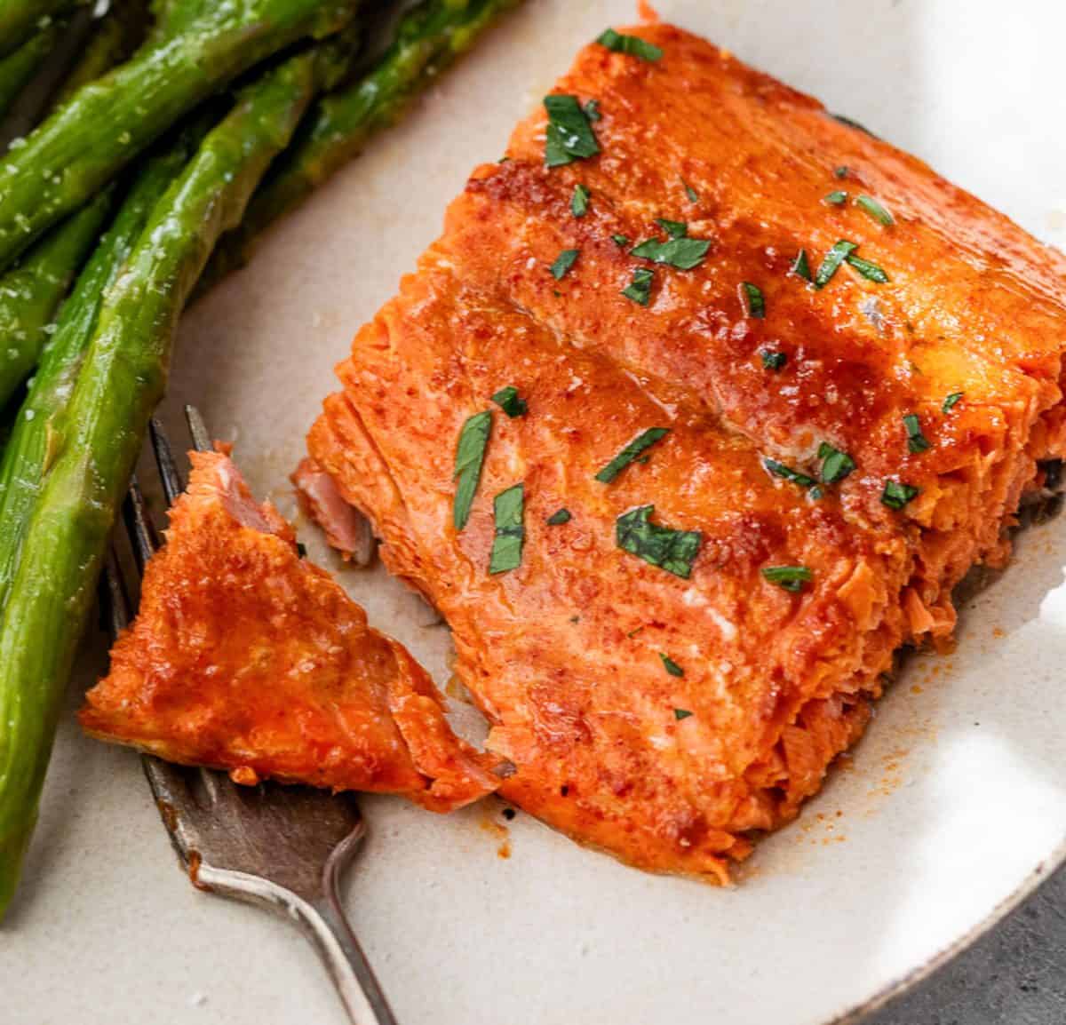 Um pedaço de salmão com mostarda e mel cozido, guarnecido com ervas, servido com aspargos em um prato branco e um garfo.