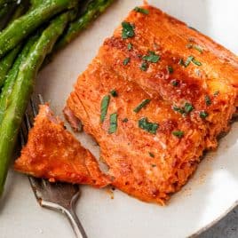 A piece of cooked honey mustard salmon garnished with herbs, served with asparagus on a white plate and a fork.