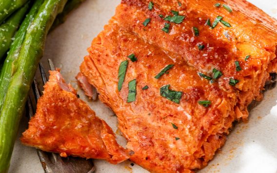 A piece of cooked honey mustard salmon garnished with herbs, served with asparagus on a white plate and a fork.