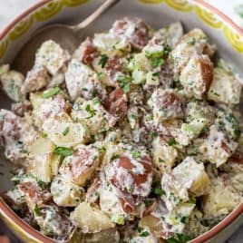 A bowl of creamy red potato salad with chopped herbs and a spoon, featuring red-skinned potatoes in a patterned bowl.