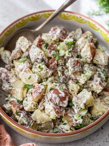 A bowl of creamy red potato salad with chopped herbs and a spoon, featuring red-skinned potatoes in a patterned bowl.