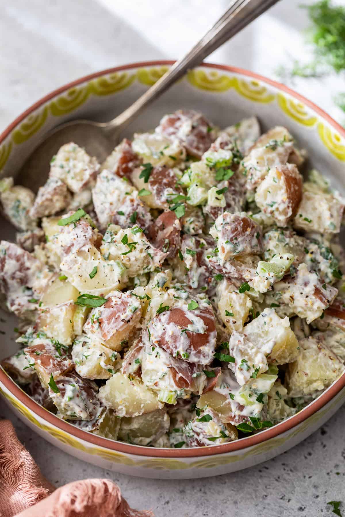 A bowl of creamy red potato salad with chopped herbs and a spoon, featuring red-skinned potatoes in a patterned bowl.