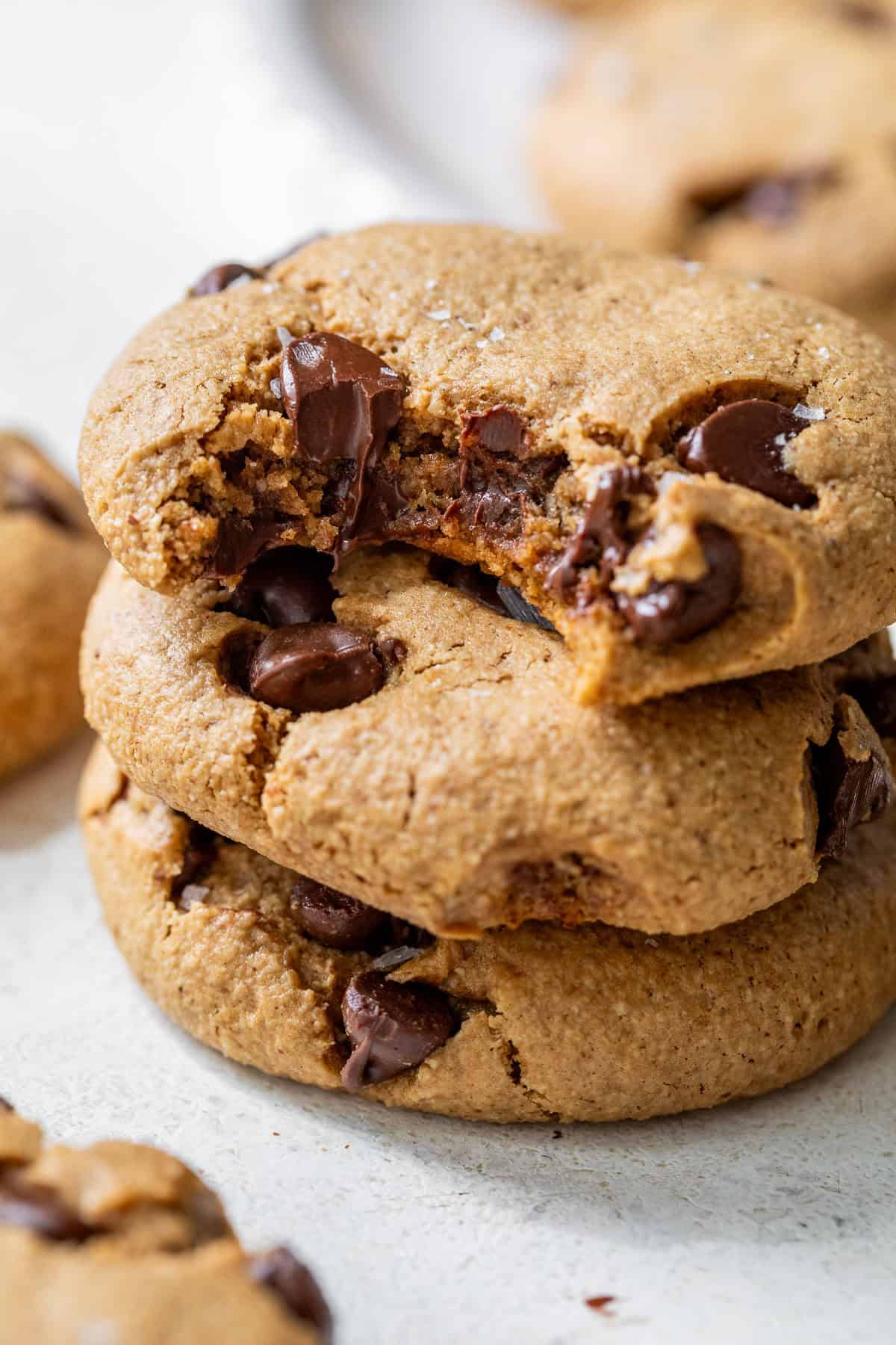 A stack of three chocolate chip almond flour cookies, with the top cookie partially bitten, on a light surface.