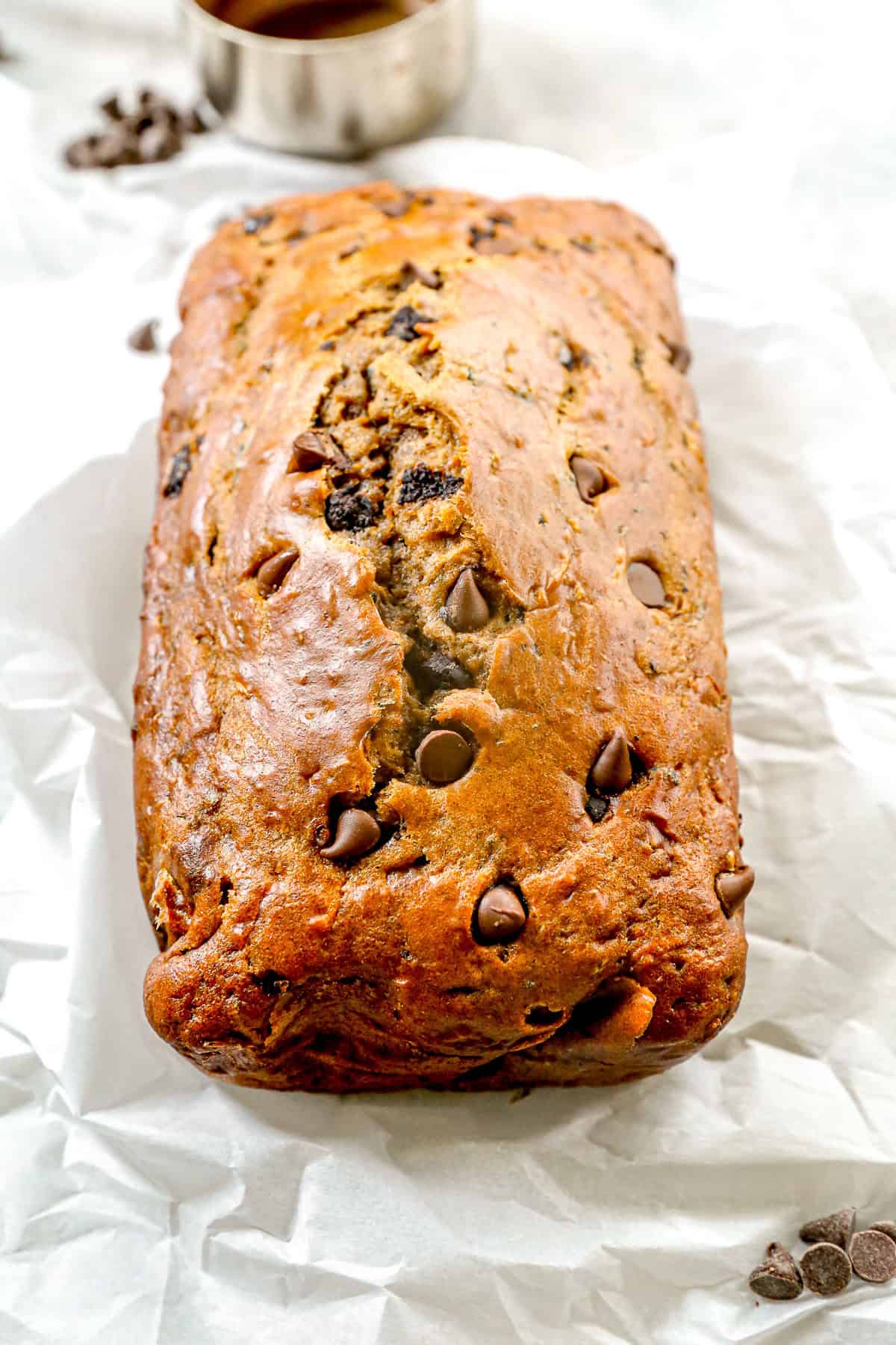 A loaf of chocolate chip zucchini bread rests on crinkled white parchment paper, surrounded by chocolate chips.
