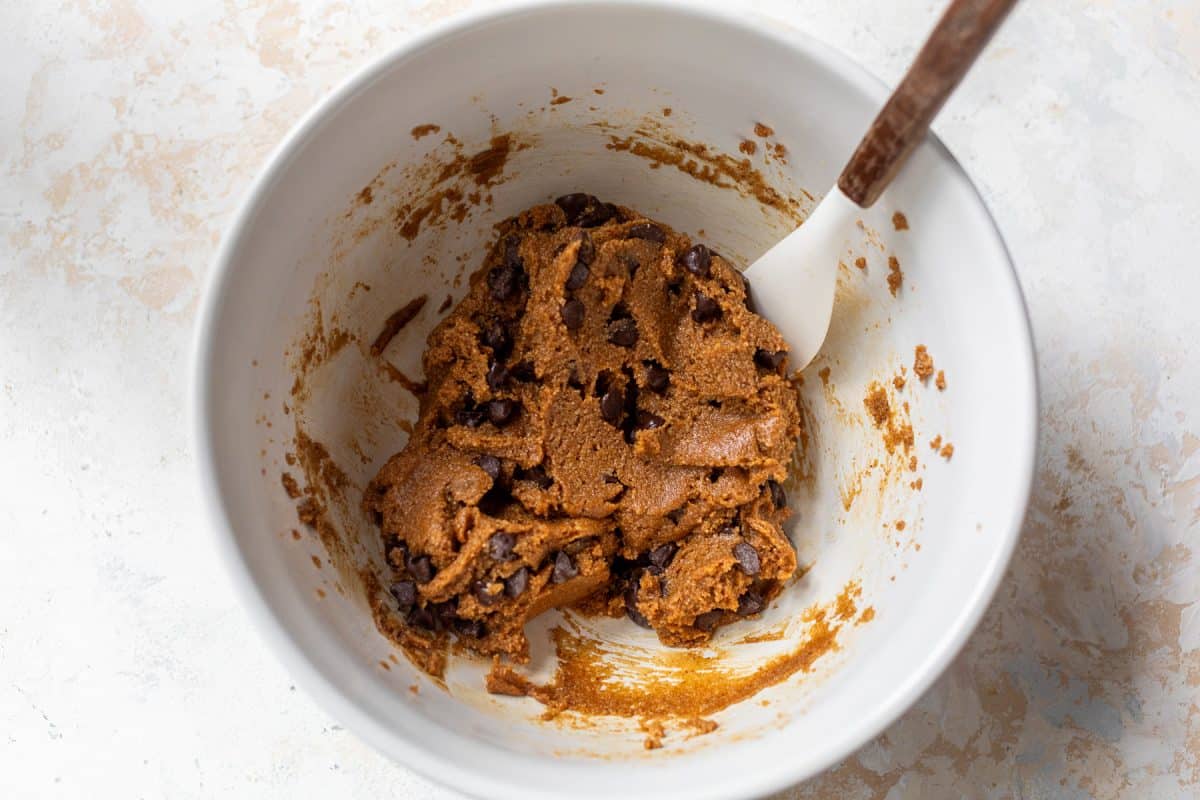 A white bowl with chocolate chip almond flour cookie dough and a white spatula on a light surface.