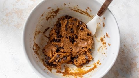 A white bowl with chocolate chip almond flour cookie dough and a white spatula on a light surface.